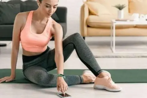 Frau in Sportkleidung sitzt auf einer Matte und bedient ihr Smartphone.