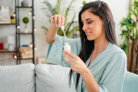 Frau mit dunklen Haaren in einem hellblauen Morgenmantel, die einen Tropfen eines Vitamin C Serums aus einer Pipette auf eine Flasche fallen lässt.