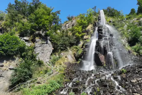 Der Trusetaler Wasserfall stürzt über schwarze Felsen in die Tiefe. Ein weißer Geländerweg führt Besucher am grünen Hang entlang.