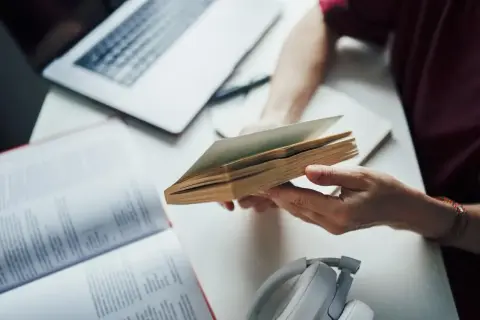 Ręce trzymają otwartą książkę, obok laptop i słuchawki. To dobry przykład, jak tworzyć bibliografię.