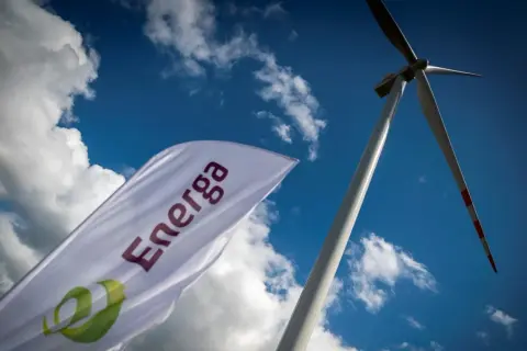 Flaga z logo Energa powiewa na tle błękitnego nieba z chmurami, obok wiatraka. Akcje Energa rosną.