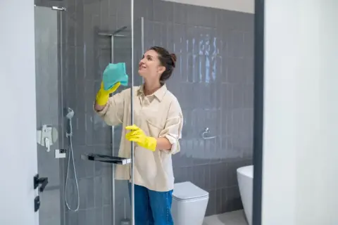 Mujer con guantes amarillos limpiando mampara ducha muy sucia con paño azul. Baño moderno con azulejos grises y sanitario blanco.
