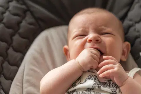 Un bebé de 3 meses no duerme, con los ojos cerrados y la mano en la boca, en una hamaca acolchada.