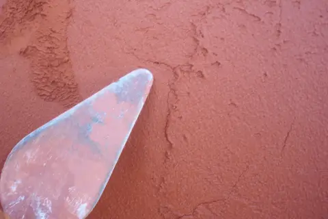 Una espátula rosa aplica mortero rojizo a una pared texturizada.