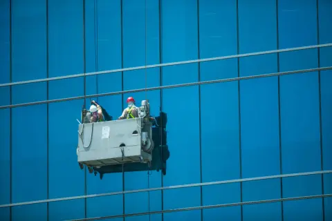 Dos trabajadores realizan limpieza en altura en una plataforma suspendida, brillando los cristales de un edificio azul.
