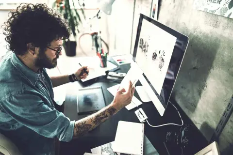 Ein Mann mit lockigen Haaren und Tattoos arbeitet an seinem Computer. Er ist im Bereich IU Kommunikationsdesign tätig und entwirft visuelle Konzepte.