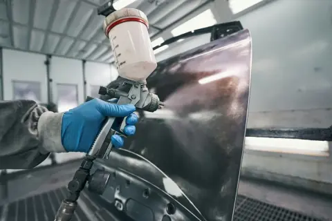 Ręka w niebieskiej rękawiczce trzyma pistolet lakierniczy, przygotowując element do malowania. Pytanie: czy na podkład epoksydowy można kłaść lakier?
