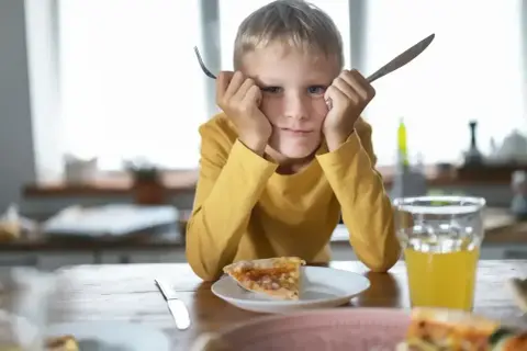 Chłopiec z widoczną wybiórczością pokarmową trzyma widelec i nóż, patrząc z niechęcią na pizzę.