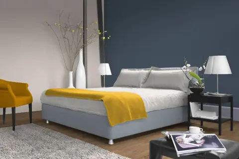 Ein Schlafzimmer mit einem Bett, das mit einer gelben Decke und Kissen in verschiedenen **blau nuancen** dekoriert ist. Ein gelber Sessel und ein Teppich ergänzen den Raum.
