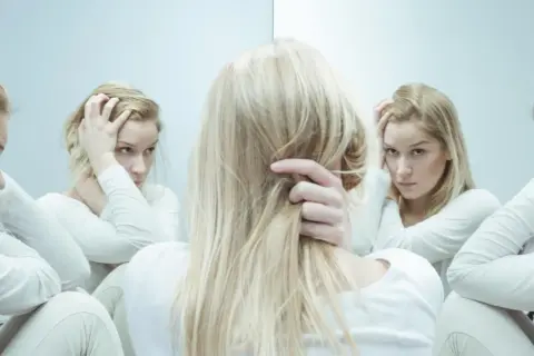 Kobieta w białej bluzie z długimi blond włosami, zmagająca się z kwetaplex, widzi swoje odbicia w lustrze, każde z innym wyrazem twarzy.
