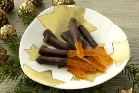 Bâtonnets d'orange confite chocolatés sur une assiette étoilée, entourés de pommes de pin dorées et de branches de sapin.