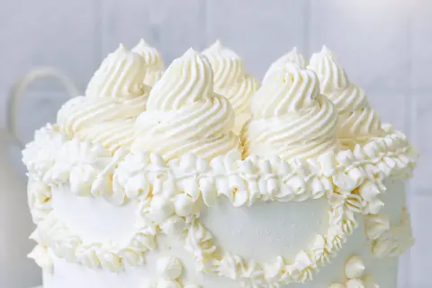 Eine üppige Buttercreme Torte mit kunstvollen Tupfen und Rüschen aus weißer Buttercreme, die auf einem weißen Hintergrund steht.