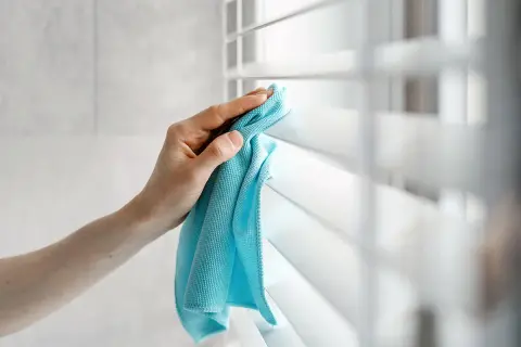 Dłoń z niebieską ściereczką czyści białe żaluzje, usuwając kurz.