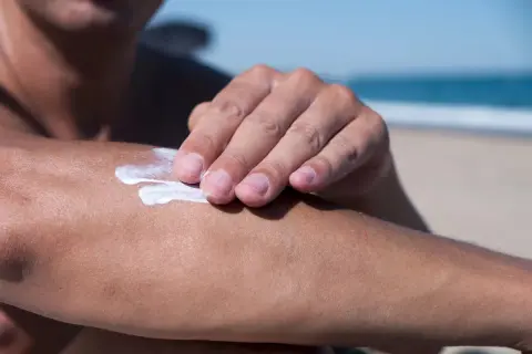 Dłoń rozsmarowuje krem z filtrem, zawierający m.in. diethylamino hydroxybenzoyl hexyl benzoate, na ramieniu na plaży.