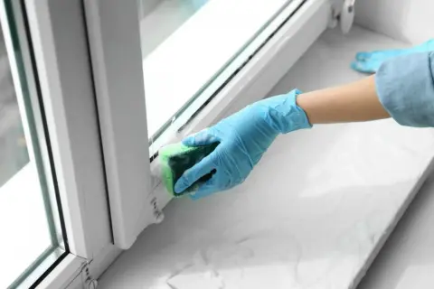 Ręka w niebieskiej rękawiczce czyści ramę okienną gąbką, usuwając klej po taśmie.
