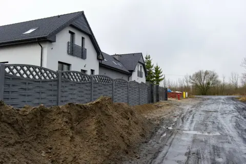 Nowy, szary płot dzieli posesje, sugerując konflikt z sąsiadem o ogrodzenie. Błotnista droga i sterta ziemi podkreślają napięcie.