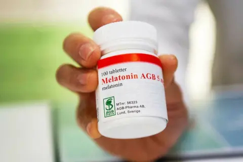 En hand håller en burk med 100 melatonin tabletter. Frågan "hur mycket melatonin kan man ta" kan besvaras genom att läsa etiketten.