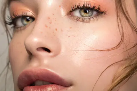 Occhi verdi con un delicato trucco lilla e pesca, labbra piene e lentiggini naturali.