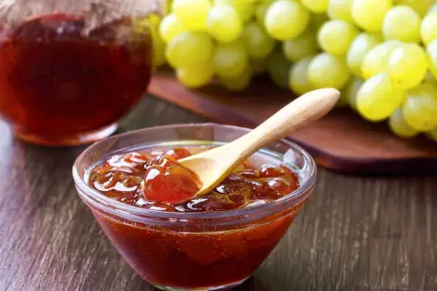 Un cuenco de cristal con mermelada de uvas casera, una cuchara de madera y un racimo de uvas verdes al fondo.