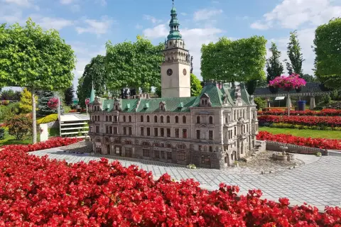 Miniatura ratusza z wieżą zegarową, otoczona czerwonymi kwiatami, w miniaturowej kotlinie kłodzkiej.