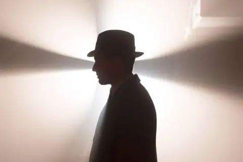 Ein Mann im Hut und Anzug, im Profil, vor einem hellen, strahlenden Hintergrund. Ein mysteriöser Blick, der an Babylon Berlin Staffel 5 erinnert.