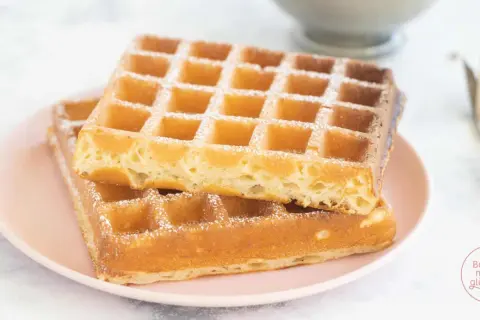 Zwei goldbraune belgische Waffeln, mit Puderzucker bestäubt, auf einem rosa Teller.