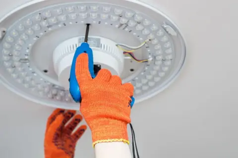 Ręka w pomarańczowej rękawicy przykręca śrubokrętem lampę sufitową LED. Dowiedz się, jak zrobić oświetlenie.