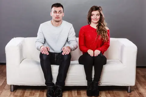 Coppia seduta su un divano bianco, lui guarda altrove con espressione incerta, lei sorride. Come superare la timidezza in amore?