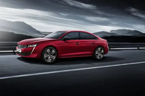 Voiture rouge Peugeot 508, un modèle à éviter, roulant sur une route avec des montagnes en arrière-plan sous un ciel dramatique.