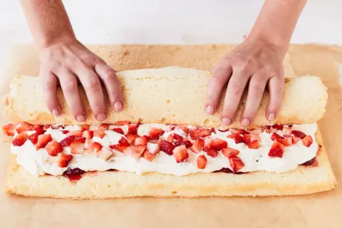 Hände rollen einen Biskuitteig mit Sahne und Erdbeeren für ein Erdbeerrolle Rezept auf.