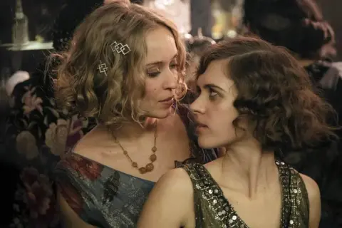 Zwei Frauen im Stil der 1920er Jahre, eine mit blonden Locken und Schmuck, die andere mit dunklen Locken, blicken sich intensiv an. Ein Moment aus Babylon Berlin Staffel 5.