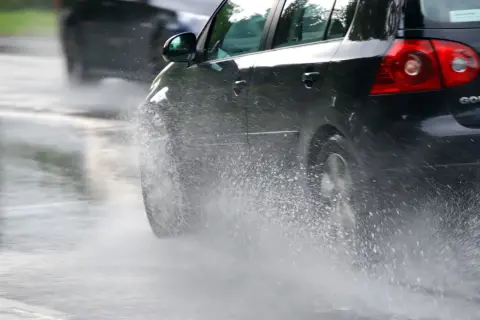 Un coche negro salpica agua al pasar por un charco. En días de lluvia, una distancia de frenado segura depende de la precaución.