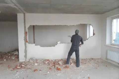Mężczyzna z młotem wyburza fragment ściany nośnej, wokół gruz.