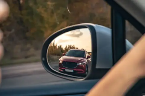 Reflejo de un coche rojo en el espejo retrovisor, preparándose para un adelantamiento. DGT.