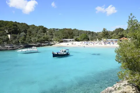 En solig dag på mallis Mallorca. Turkosblått vatten, en vit yacht och en blåvit båt i en vik med en livlig strand.