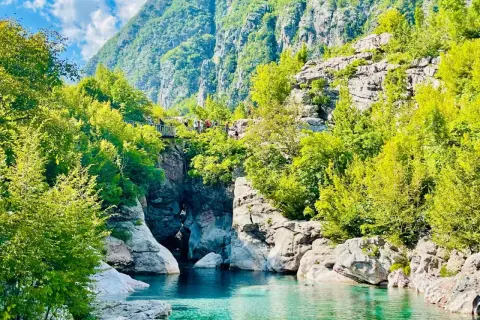 Aguas turquesas fluyen entre rocas y vegetación exuberante, bajo imponentes montañas en Albania. Un puente peatonal cruza el cañón, invitando a explorar las **montañas malditas de Albania**.