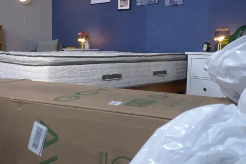 A mattress that expands when opened is shown, partially covered by a cardboard box and plastic packaging.