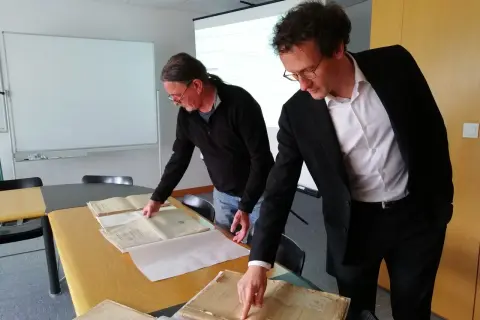 Deux hommes consultent des registres d'état civil aux archives départementales du Morbihan. L'un pointe un document ancien.