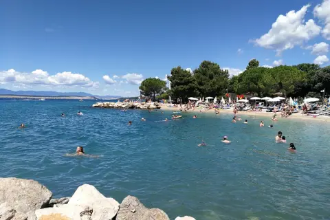 Crikvenica atrakcje: plaża pełna ludzi kąpiących się i opalających, błękitne morze, zielone drzewa i góry w tle.
