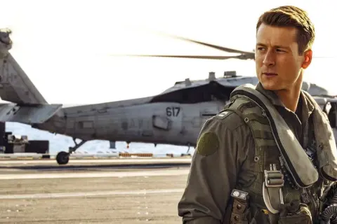 Glen Powell w filmach jako pilot na pokładzie lotniskowca, z helikopterem w tle.