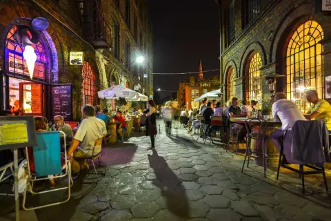 Wieczór na ul. Piotrkowskiej w Łodzi. Ludzie siedzą przy stolikach w restauracjach, ciesząc się atmosferą.