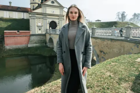 Kobieta w szarym płaszczu i golfie, idealna stylizacja na 14 stopni. W tle zamek i fosą.