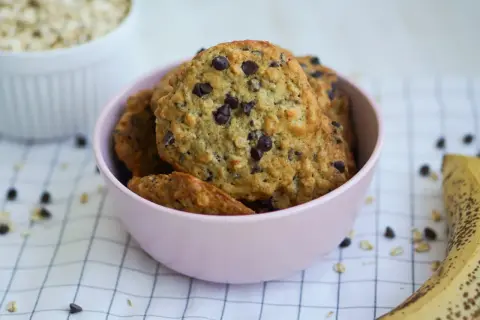 Ciasteczka bez cukru z kawałkami czekolady w różowej miseczce. Obok leży banan i miska z płatkami owsianymi. Idealne przepisy dla dbających o zdrowie.