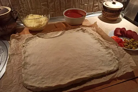 Gotowe ciasto na pizzę czeka na dodatki: ser, sos, salami, papryka i oliwki.