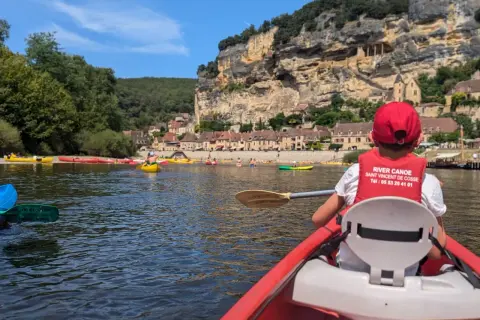 Enfant en canoë rouge, gilet de sauvetage "River Canoe", pour une visite insolite autour de Sarlat, avec des falaises et des maisons troglodytes en arrière-plan.