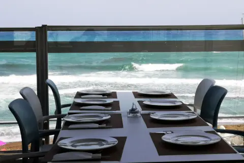 Mesa puesta en el chiringuito la sal, con platos y cubiertos, frente a un mar turquesa y olas rompiendo.