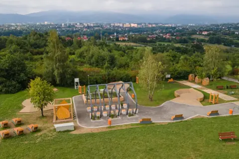 Nowoczesny plac zabaw i siłownia plenerowa, otoczone zielenią, z widokiem na miasto. W oddali widać trzy lipki.