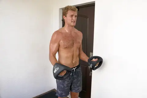 A muscular man with sweaty boxer forearms holds two black dumbbells, ready for a workout.