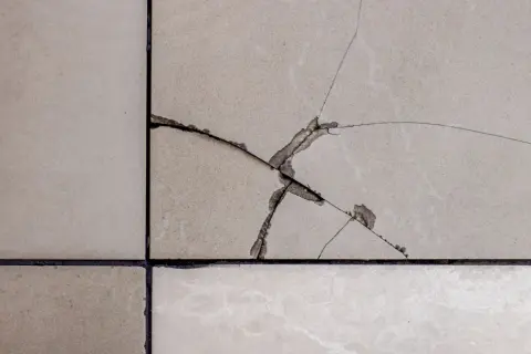 Gros plan sur un carrelage beige avec une fissure traversant plusieurs carreaux, révélant la couche inférieure.