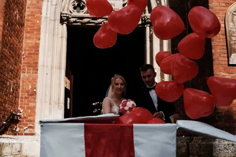 Para młoda wyskakuje z pudełka wśród czerwonych serduszek. Śmieszna niespodzianka dla pary młodej przed kościołem.